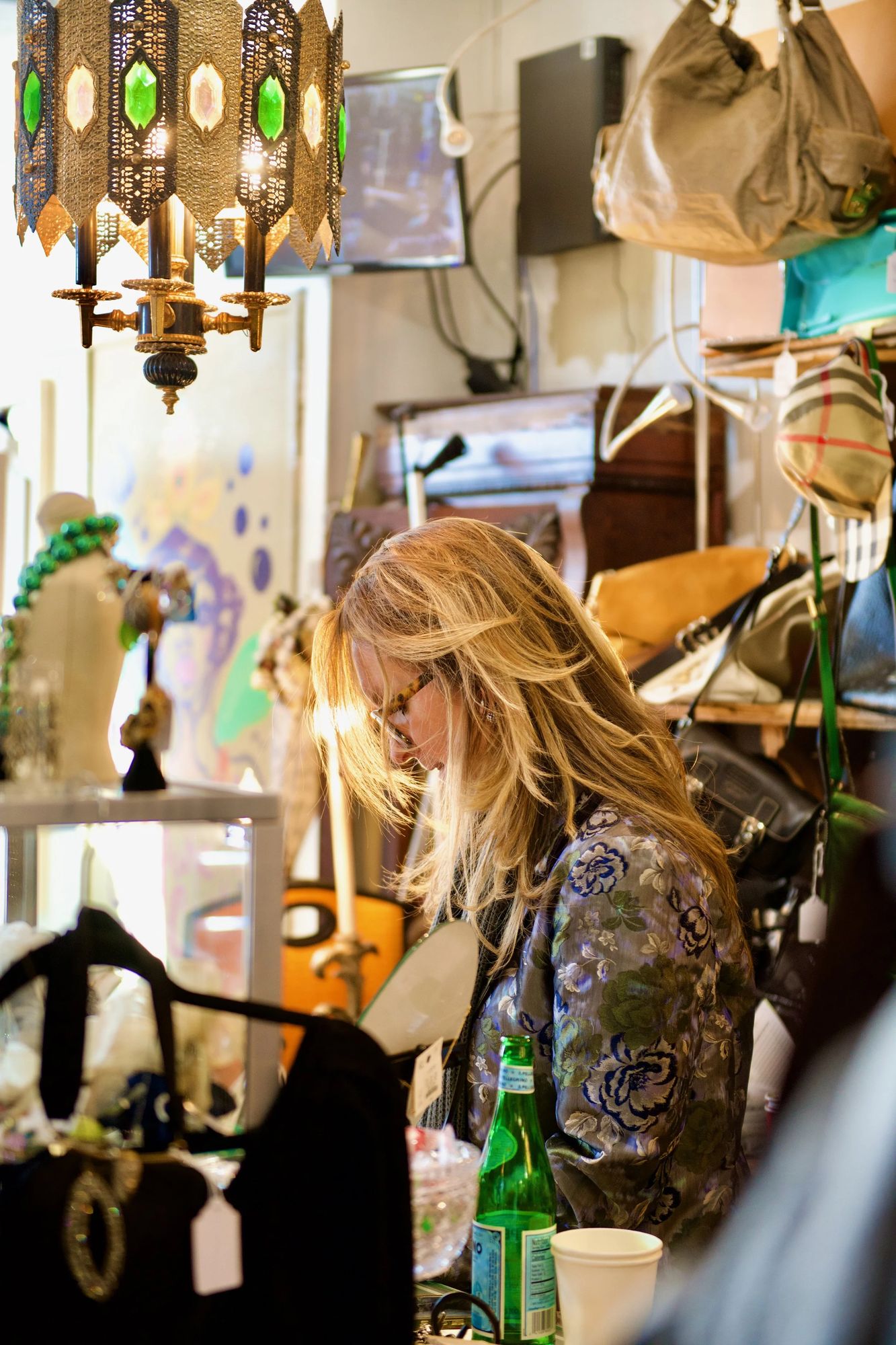 Denise at the counter beneath a jeweled pendant lamp, surrounded by handbags