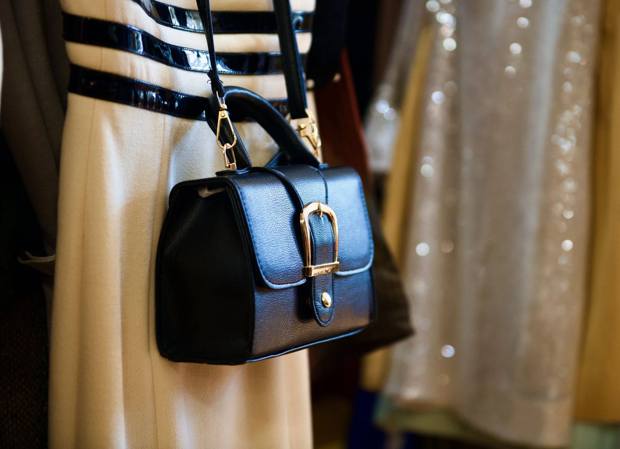 A structured black handbag with a gold buckle, hanging against a striped coat beside a sequined dress